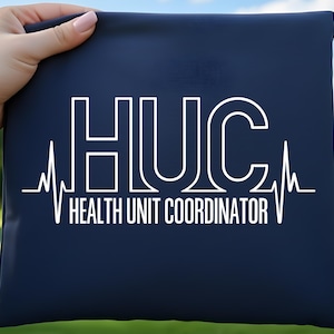 Peut inclure: Un objet rectangulaire bleu marine avec le texte blanc "HUC HEALTH UNIT COORDINATOR" et un graphique de ligne de battement de cœur blanc. L'objet est tenu par une main avec un arrière-plan vert flou.