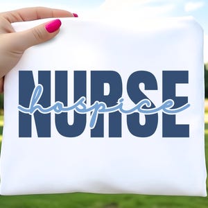 May include: White t-shirt with the word "NURSE" in dark blue block letters and the word "hospice" in light blue script. The shirt is held up against a green background.