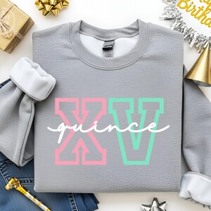 Puede incluir: Una sudadera gris con el texto "XV quince" en letras de bloque rosas y verdes y cursiva blanca. La sudadera tiene pu&ntilde;os blancos y est&aacute; rodeada de decoraciones de cumplea&ntilde;os, incluyendo una caja de regalo dorada y una pancarta de "Happy Birthday".