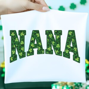 Könnte beinhalten: Weißes T-Shirt mit dem Wort "NANA" in großen, grünen Buchstaben mit Kleeblattmuster und schwarzer Umrandung. Das T-Shirt wird von einer Person hochgehalten.