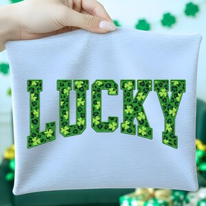 May include: A pale blue t-shirt featuring the word "LUCKY" in large, green, block letters. The letters have a leopard print pattern and shamrock accents, ideal for St. Patrick's Day celebrations.