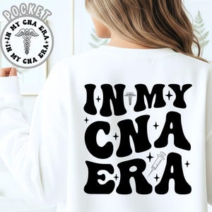 Puede incluir: Sudadera blanca con el texto "IN MY CNA ERA" en letras negras grandes de estilo retro en la espalda. El diseño incluye un símbolo médico, estrellas y una jeringa. Un diseño más pequeño está en la parte delantera.