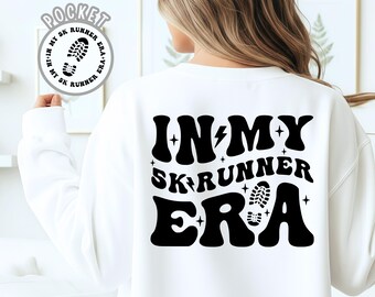 Op de afbeelding: Witte sweatshirt met de zwarte tekst "IN MY SK RUNNER ERA" in retro-stijl. Het ontwerp bevat een bliksemschicht, sterren en een voetafdruk. Een cirkelvormig logo met tekst en voetafdruk is zichtbaar.