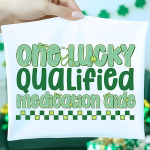 May include: White pillowcase with the text "one lucky qualified medication aide" in shades of green. The design includes a stethoscope, shamrocks, and a checkered border. The pillowcase is held up by a hand.