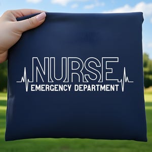 Puede incluir: Una bolsa tote azul marino con las palabras "NURSE EMERGENCY DEPARTMENT" en blanco, junto con un gráfico de línea de latido cardíaco blanco. La bolsa se sostiene sobre un fondo de hierba verde y árboles.