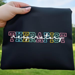 May include: A black pouch featuring the text "RESPIRATORY THERAPIST". The word "THERAPIST" is in a block letter style with pink, blue, and yellow outlines, and "RESPIRATORY" is in a cursive font.
