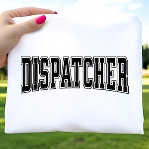 May include: A white t-shirt with the word "DISPATCHER" printed in large, bold, black letters with a white outline. The text is arched across the shirt. The shirt is held up by a hand with pink nail polish.