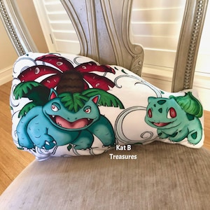 May include: A white pillow featuring a colorful illustration of two Pokémon characters, Venusaur and Bulbasaur. The Venusaur is a large, blue and green creature with a red flower on its back. Bulbasaur is smaller, green, and smiling. The pillow is on a chair.