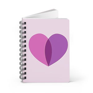 May include: A spiral-bound notebook with a light pink cover featuring a heart design. The heart is composed of two overlapping halves, one pink and one purple. The notebook's pages are white.