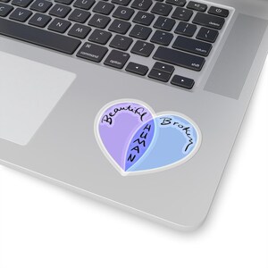 May include: A sticker with a heart shape divided into two overlapping sections. The left section is purple and says "Beautiful" and the right section is blue and says "Broken". The overlapping section says "Human" in black text.