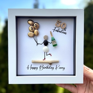 May include: Framed pebble art with a white border, depicting a figure with gold balloons and a champagne bottle. The text reads "50 & Fabulous" and "Happy Birthday Kerry". The figure is wearing a white skirt and red heels. The artwork is a birthday theme.