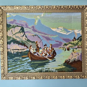 May include: A framed needlepoint picture depicting a group of people in a boat on a lake. The lake is surrounded by mountains and trees. The boat is brown and has a white stripe. The people in the boat are wearing colorful clothing.