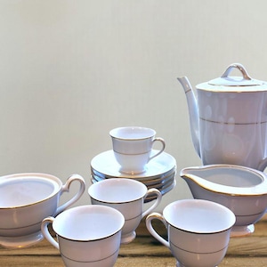 May include: A white porcelain tea set with gold trim, including a teapot, creamer, sugar bowl, cups, and saucers. The set is arranged on a wooden surface, showcasing a classic and elegant design.
