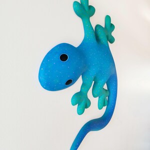Puede incluir: Una escultura caprichosa de un lagarto azul con un patr&oacute;n de lunares y una cola curvada. El lagarto tiene una cabeza redondeada con dos ojos negros y est&aacute; posado como si estuviera trepando. La escultura es una vibrante obra de arte mural.