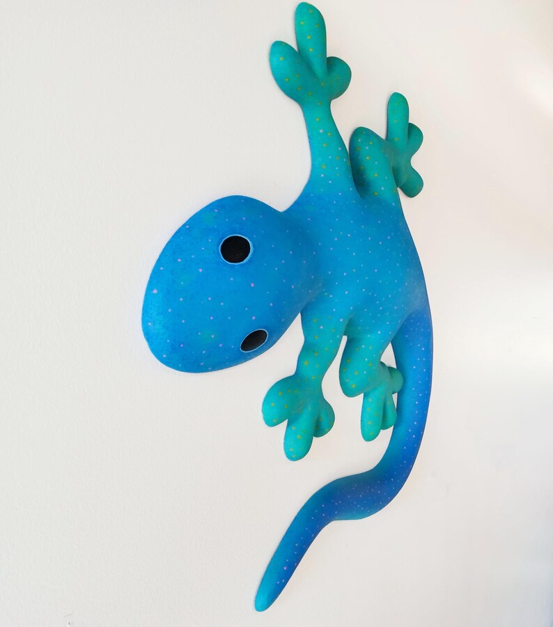 Puede incluir: Una escultura caprichosa de un lagarto azul con un patr&oacute;n de lunares y una cola curvada. El lagarto tiene una cabeza redondeada con dos ojos negros y est&aacute; posado como si estuviera trepando. La escultura es una vibrante obra de arte mural.