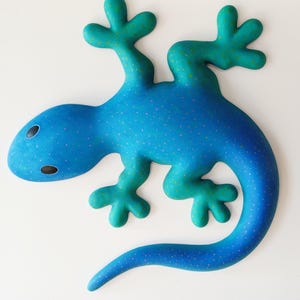 Puede incluir: Escultura de lagarto estilizada en tonos azul y turquesa. El lagarto presenta un patr&oacute;n moteado y formas redondeadas. Es probable que la obra de arte sea una pieza decorativa para una pared o una mesa.