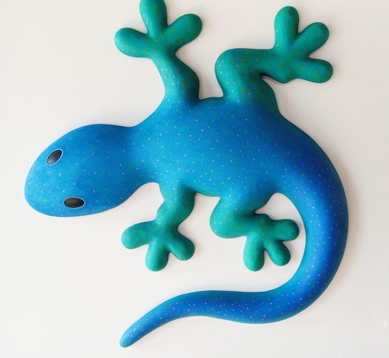 Puede incluir: Escultura de lagarto estilizada en tonos azul y turquesa. El lagarto presenta un patr&oacute;n moteado y formas redondeadas. Es probable que la obra de arte sea una pieza decorativa para una pared o una mesa.