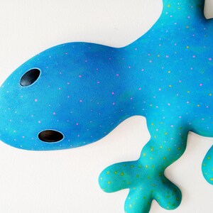 Puede incluir: Una escultura abstracta de lagarto en tonos azules y turquesas. El lagarto tiene una cabeza redondeada con dos agujeros negros para los ojos y est&aacute; decorado con puntos rosas y amarillos. La obra de arte est&aacute; sobre un fondo blanco.