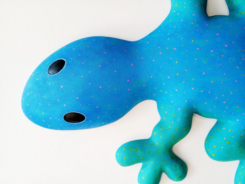 Puede incluir: Una escultura abstracta de lagarto en tonos azules y turquesas. El lagarto tiene una cabeza redondeada con dos agujeros negros para los ojos y est&aacute; decorado con puntos rosas y amarillos. La obra de arte est&aacute; sobre un fondo blanco.