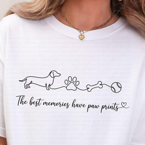 May include: A white short-sleeved t-shirt featuring a black continuous line drawing of a dachshund, a paw print, a bone, and a ball. Below the graphic, the text reads, "The best memories have paw prints" followed by a small heart symbol.