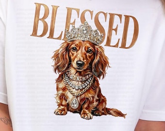 May include: White t-shirt with the word "BLESSED" in gold lettering. The shirt features a detailed illustration of a dachshund wearing a crown and jewelry. The dachshund is brown with long, flowing hair.