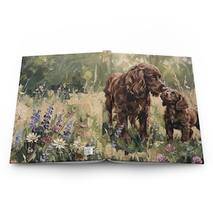 Puede incluir: Diario de tapa dura con una pintura al óleo de dos perros marrones en un campo de flores silvestres. Los perros se representan nariz con nariz en un prado cubierto de hierba con flores moradas, blancas y rosas. El diario está abierto.