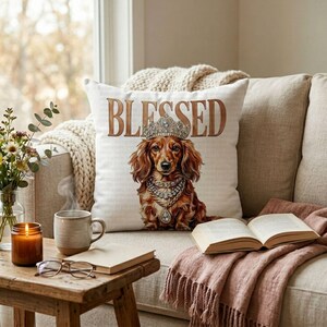 May include: A decorative throw pillow with the word "BLESSED" in brown lettering. The pillow features a regal dachshund wearing a crown and jewelry. The scene includes a coffee table with a book, glasses, and a candle.