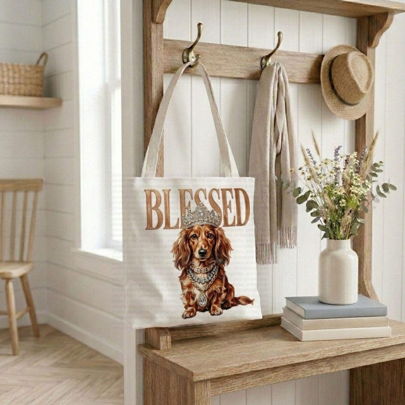 May include: A cream-colored tote bag with the word "BLESSED" in gold lettering. The bag features a regal dachshund illustration wearing a crown and jewelry. The bag hangs on a wooden coat rack with a hat and scarf.