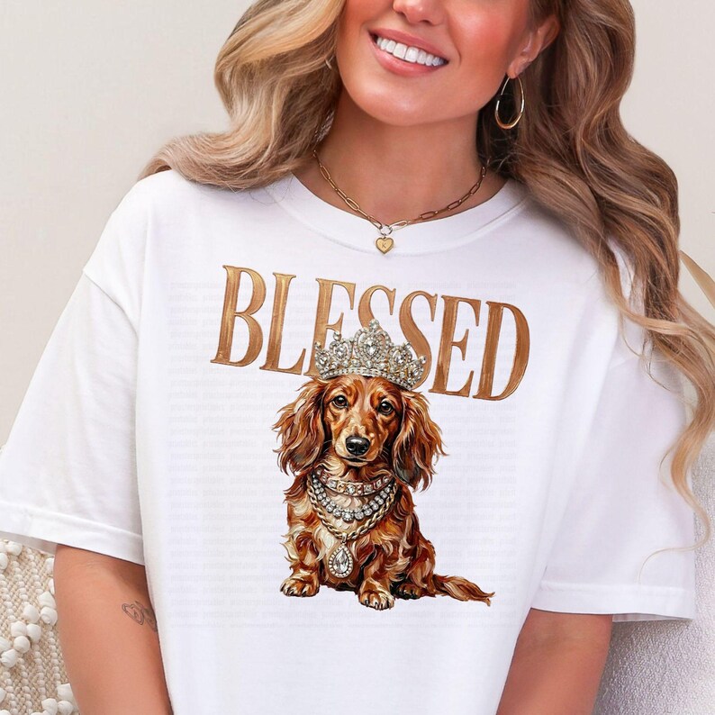 May include: White t-shirt with the word "BLESSED" in gold lettering. The shirt features a detailed illustration of a dachshund wearing a crown and jewelry. The dachshund is brown with long, flowing hair.