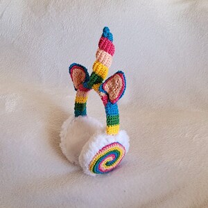 May include: Rainbow crochet ear warmers with a unicorn horn detail. The ear warmers are white and fluffy, with a colorful crocheted design. The unicorn horn is made of crocheted yarn in shades of pink, yellow, blue, and green.