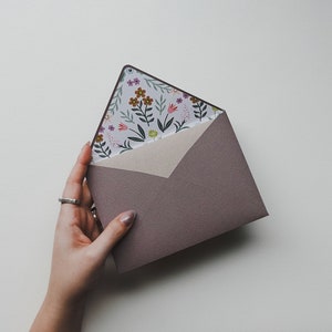 K&ouml;nnte beinhalten: Ein lila Umschlag mit einem Blumenmuster auf der Innenseite. Der Umschlag wird von einer Hand mit einem silbernen Ring gehalten.