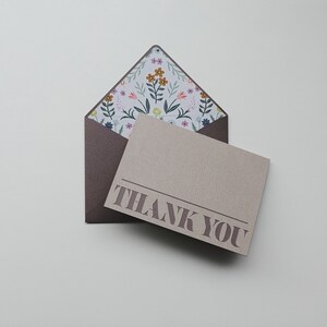 K&ouml;nnte beinhalten: Ein brauner Umschlag mit einer floralen Musterung und einer beigefarbenen Karte mit dem Text "THANK YOU" in brauner Farbe.