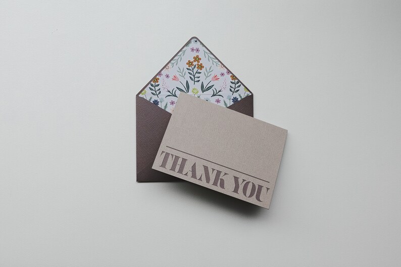 K&ouml;nnte beinhalten: Ein brauner Umschlag mit einer floralen Musterung und einer beigefarbenen Karte mit dem Text "THANK YOU" in brauner Farbe.