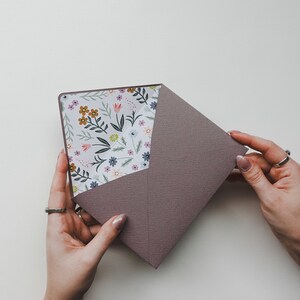 K&ouml;nnte beinhalten: Ein lila Umschlag mit einer floralen Musterung, die das Innere auskleidet. Der Umschlag wird von zwei H&auml;nden offen gehalten.