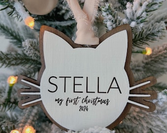 Könnte beinhalten: Weißer, katzenförmiger Weihnachtsbaumschmuck aus Holz mit dem Namen Stella, dem Text "my first christmas 2024" und einem braunen Band.