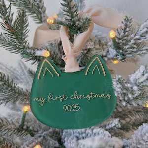 May include: A green cat-shaped Christmas ornament with gold accents and the text "my first christmas 2025". The ornament hangs from a cream-colored ribbon and is displayed on a snow-covered Christmas tree.