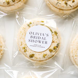 May include: A collection of vanilla shower steamers, individually wrapped in clear plastic. The round steamers are a light beige colour, dotted with dried herbs, and have a white label with the text "Olivia's Bridal Shower".