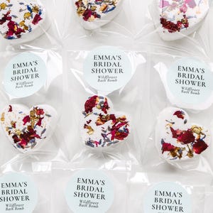 Peut inclure: Bombes de bain en forme de cœur, décorées de pétales de fleurs séchées rouges, blanches et jaunes, sont emballées dans du plastique transparent. Chaque paquet porte un autocollant rond avec "EMMA'S BRIDAL SHOWER Wildflower Bath Bomb."
