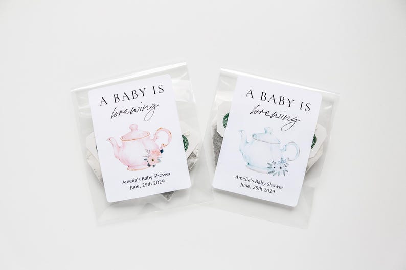 May include: Two clear plastic pouches containing tea bags, each with a white label featuring the text A BABY IS brewing and Amelia's Baby Shower June, 29th 2029. One label shows a pink teapot illustration, the other a light blue teapot illustration.