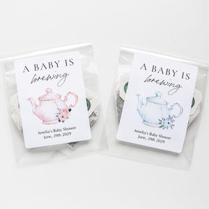 May include: Two clear plastic bags containing tea, each with a white label featuring the text A BABY IS brewing, Amelia's Baby Shower, June, 29th 2029. One label shows a pink teapot with flowers, the other a light blue teapot with flowers.