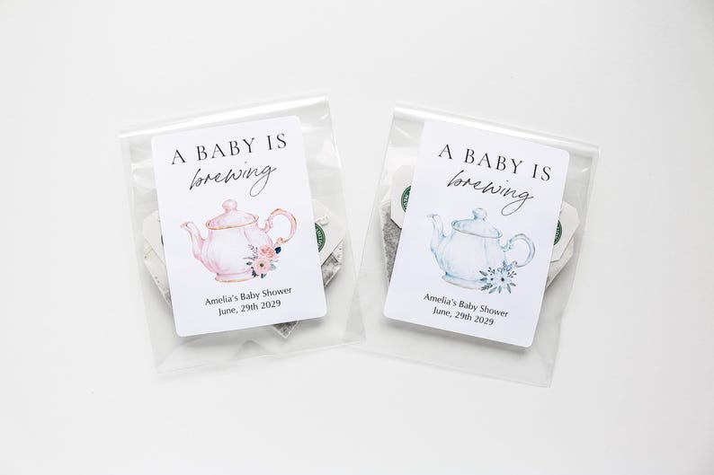 May include: Two clear plastic bags containing tea, each with a white label featuring the text A BABY IS brewing, Amelia's Baby Shower, June, 29th 2029. One label shows a pink teapot with flowers, the other a light blue teapot with flowers.