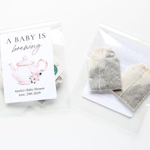 May include: Two clear plastic pouches containing tea bags. The left pouch features a white label with a pink teapot illustration, floral accents, and the text A BABY IS brewing Amelia's Baby Shower June, 29th 2029. The right pouch shows two tea bags.