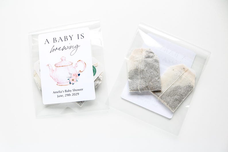 May include: Two clear plastic pouches containing tea bags. The left pouch features a white label with a pink teapot illustration, floral accents, and the text A BABY IS brewing Amelia's Baby Shower June, 29th 2029. The right pouch shows two tea bags.
