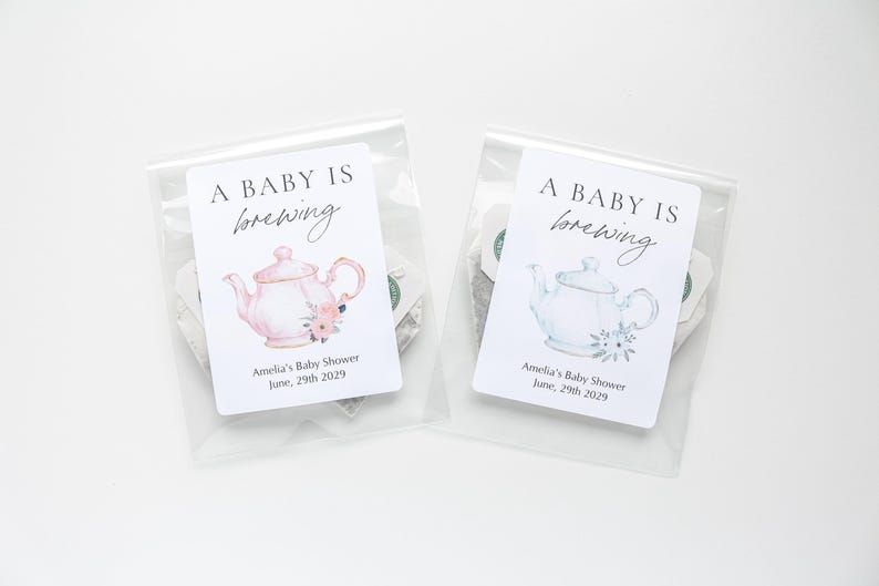 May include: Two clear plastic pouches containing tea bags, each with a white label featuring the text A BABY IS brewing, Amelia's Baby Shower, and June, 29th 2029. One label displays a pink teapot with flowers, the other a blue teapot with flowers.