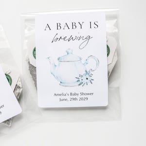 May include: A clear plastic packet containing a tea bag with a white rectangular tag. The tag features a watercolor illustration of a light blue teapot with flowers, the text A BABY IS brewing, and Amelia's Baby Shower June, 29th 2029.