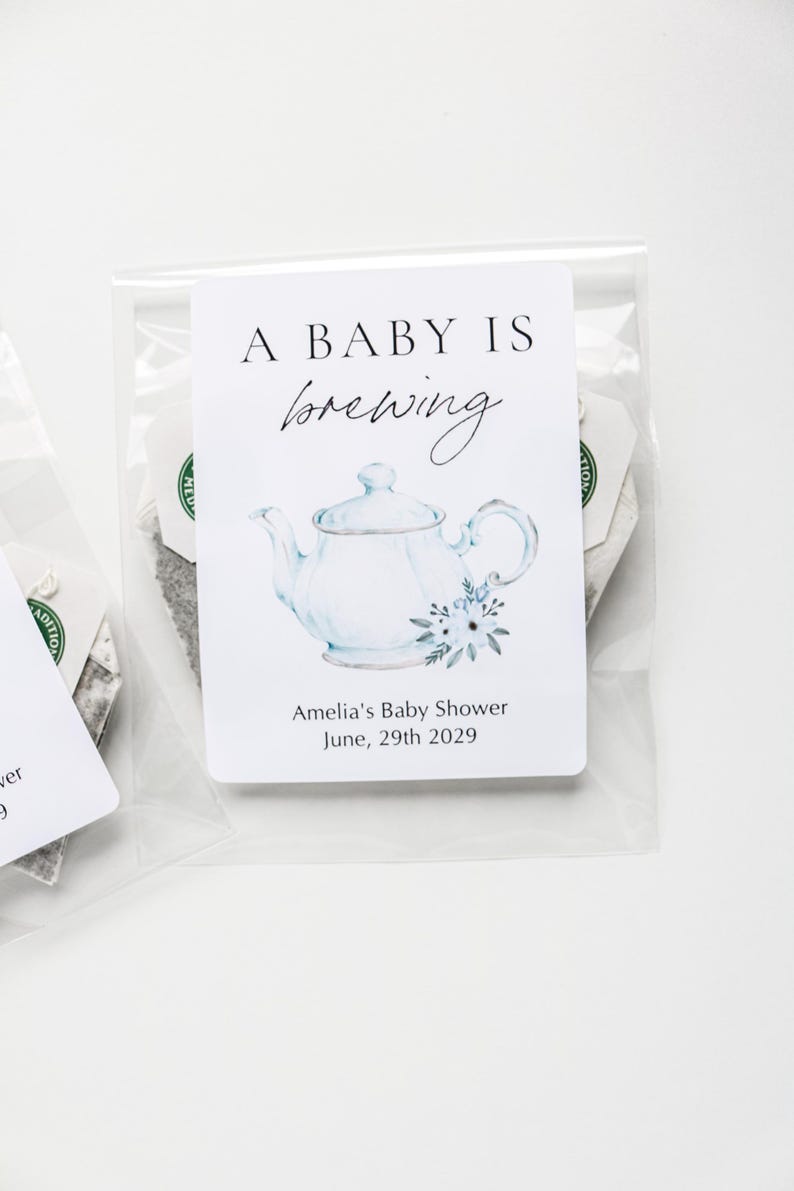 May include: A clear plastic packet containing a tea bag with a white rectangular tag. The tag features a watercolor illustration of a light blue teapot with flowers, the text A BABY IS brewing, and Amelia's Baby Shower June, 29th 2029.