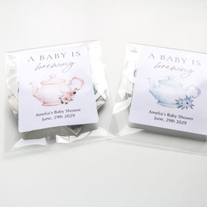 May include: Two clear plastic bags containing tea favors, each with a white label reading A BABY IS brewing, Amelia's Baby Shower, June, 29th 2029. The left label features a pink teapot illustration, while the right label features a blue teapot illustration.