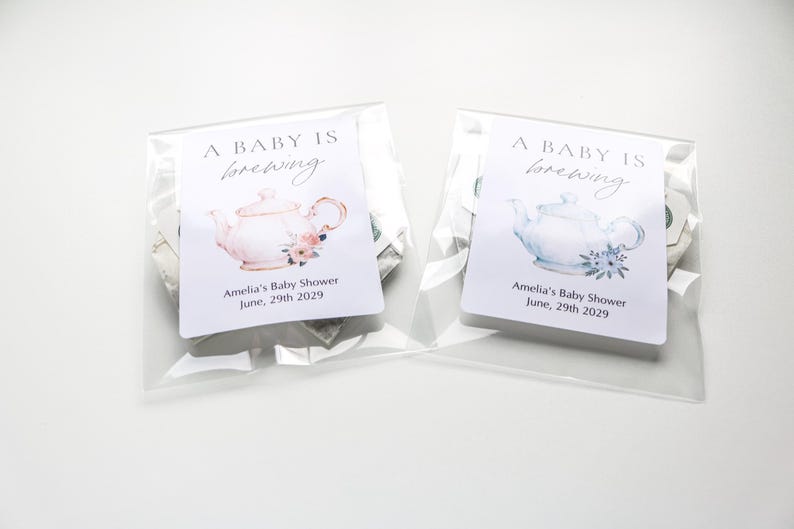 May include: Two clear plastic bags containing tea favors, each with a white label reading A BABY IS brewing, Amelia's Baby Shower, June, 29th 2029. The left label features a pink teapot illustration, while the right label features a blue teapot illustration.