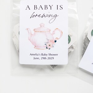 May include: A clear plastic bag containing a tea bag with a white rectangular label. The label features a pink teapot illustration with floral accents, the text A BABY IS brewing, and the text Amelia's Baby Shower June, 29th 2029.