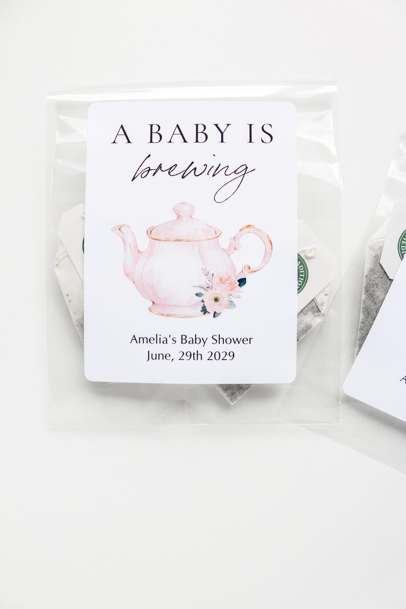 May include: A clear plastic bag containing a tea bag with a white rectangular label. The label features a pink teapot illustration with floral accents, the text A BABY IS brewing, and the text Amelia's Baby Shower June, 29th 2029.